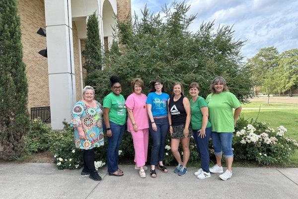 Six teachers posing for a picture with Dr. Weber in front of flowers and shrubbery.